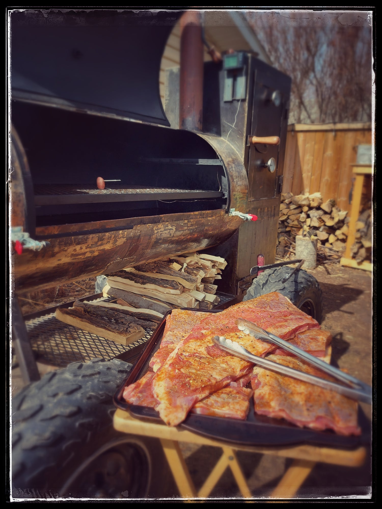 A stack of pork side ribs being readied to put on the wood fired smoker seen in the background.